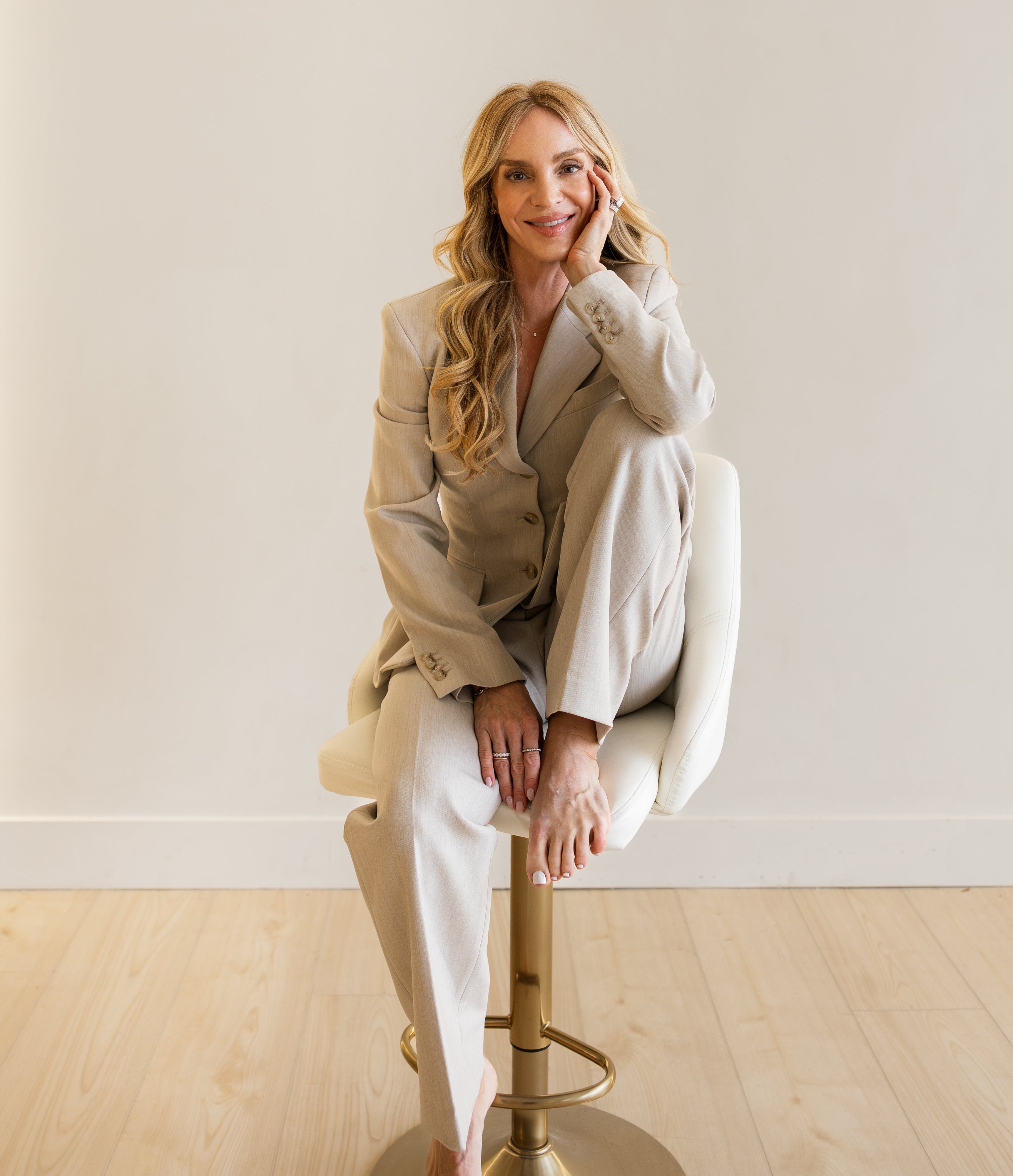 Woman in a beige suit sitting on a white chair with a gold base against a plain wall.